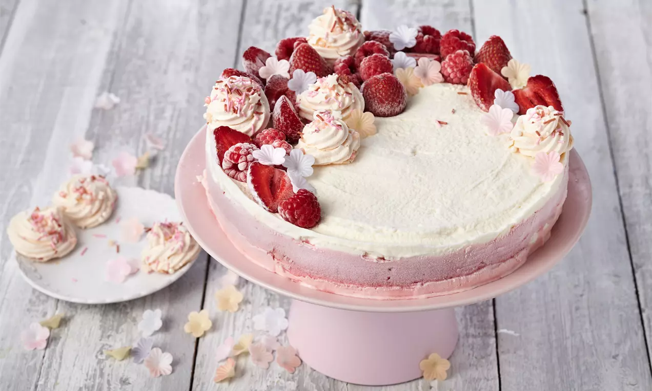 Eistorte mit weißer Sahnecreme, rosa Schichten, frischen Himbeeren und Erdbeeren, dekoriert mit Baiser und Blüten auf rosa Tortenständer