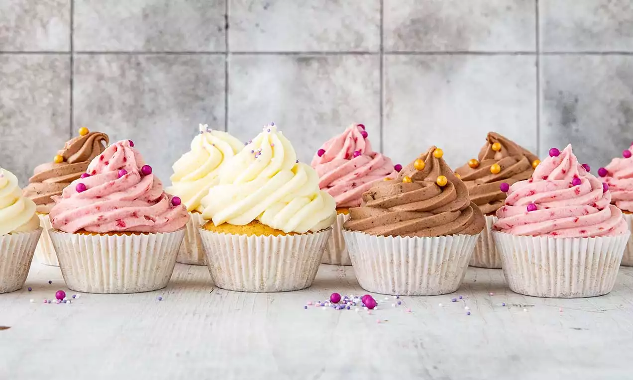 Vanille-Cupcakes in Papierförmchen mit fluffigen Toppings aus weißer Creme, rosa Frosting und schokoladig brauner Creme, bestreut mit bunten Perlen