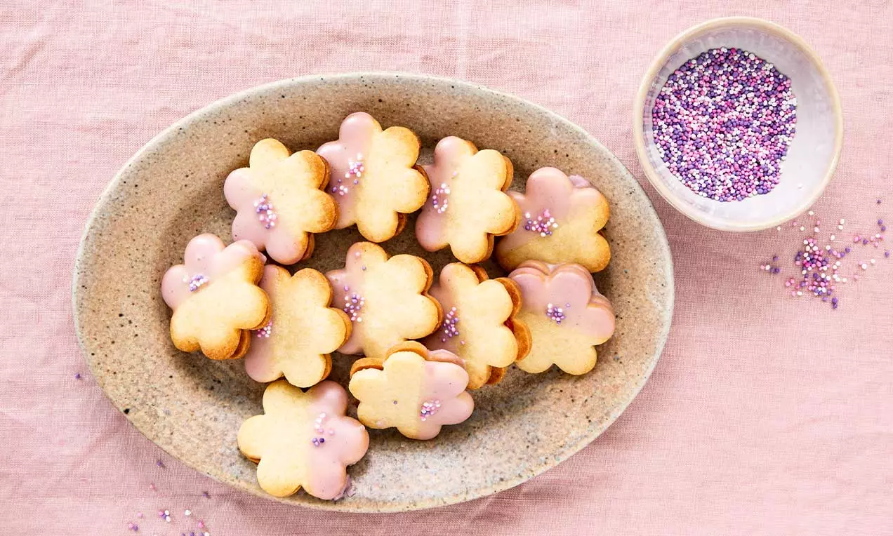 Blütenförmige, goldgelbe Guetzli mit rosa Glasur und lilafarbenen Zuckerperlen auf einem bogenförmigen Keramikteller auf rosafarbenem Stoff
