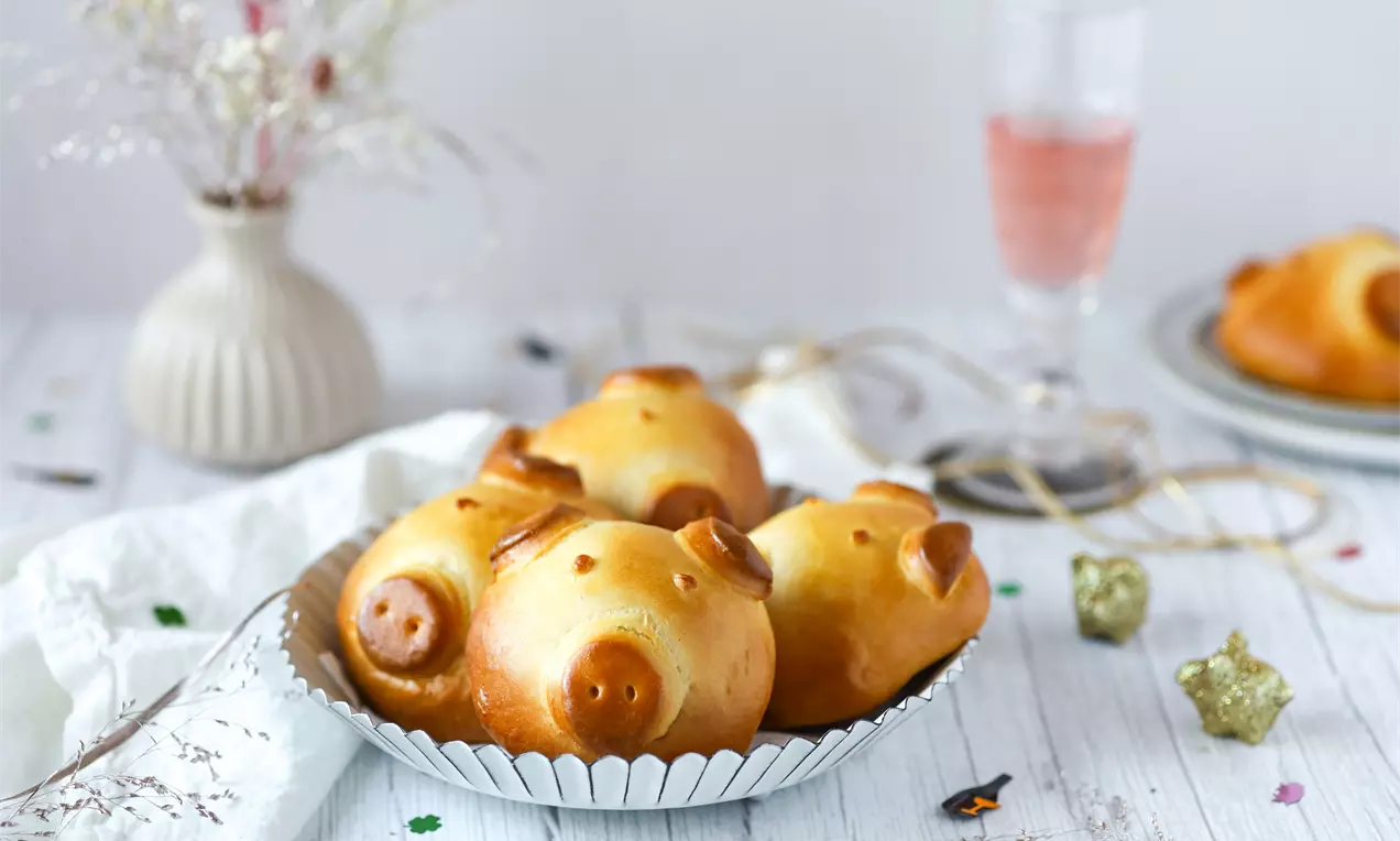 Goldbraune, glänzende Glücksschweinchen aus Hefeteig mit zarten Details in einer silbernen Schale auf weißem Tisch mit festlicher Dekoration