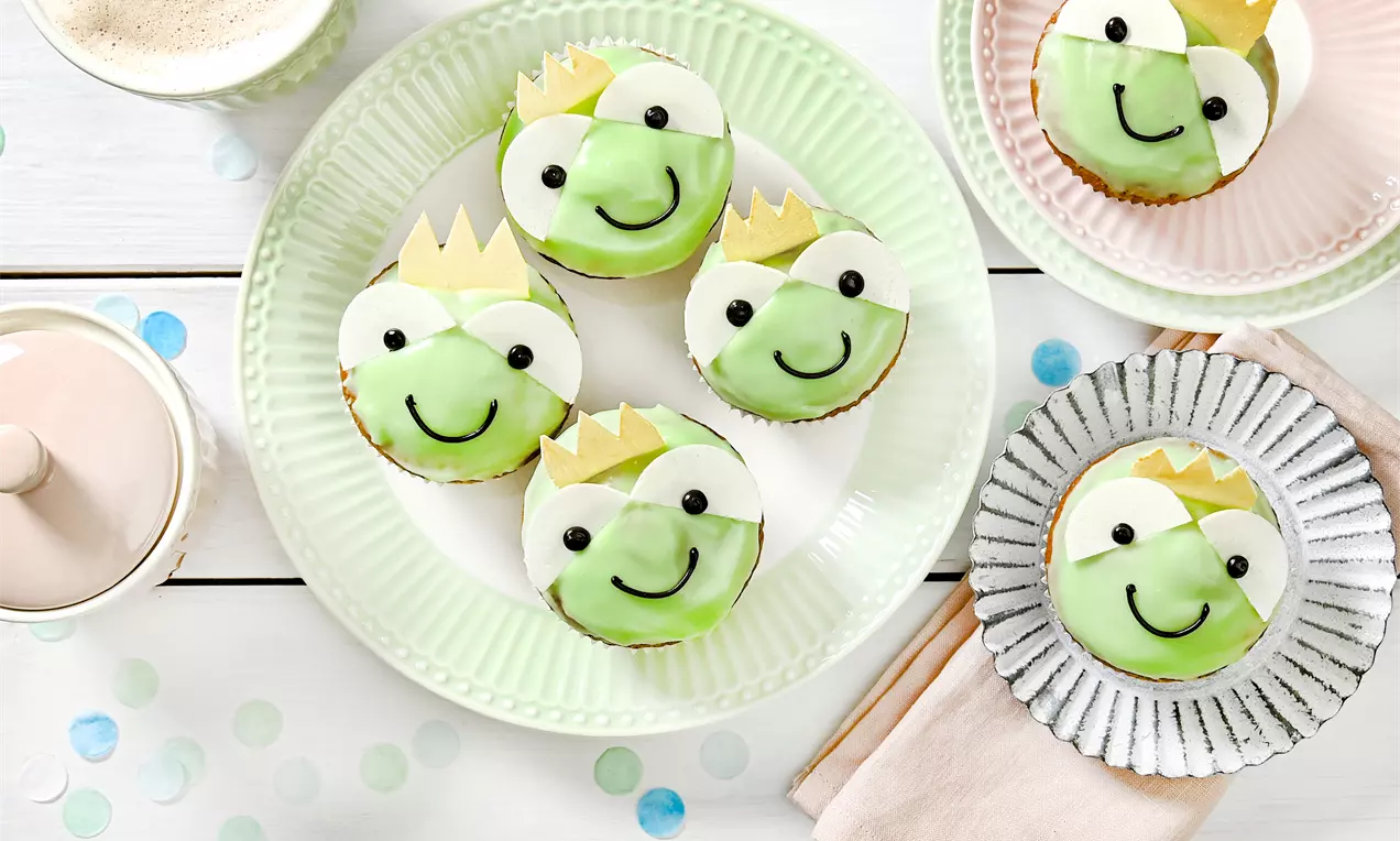 Muffins décorés de visages de grenouilles vertes avec couronnes dorées en pâte, posés sur des assiettes pastel entourées de confettis