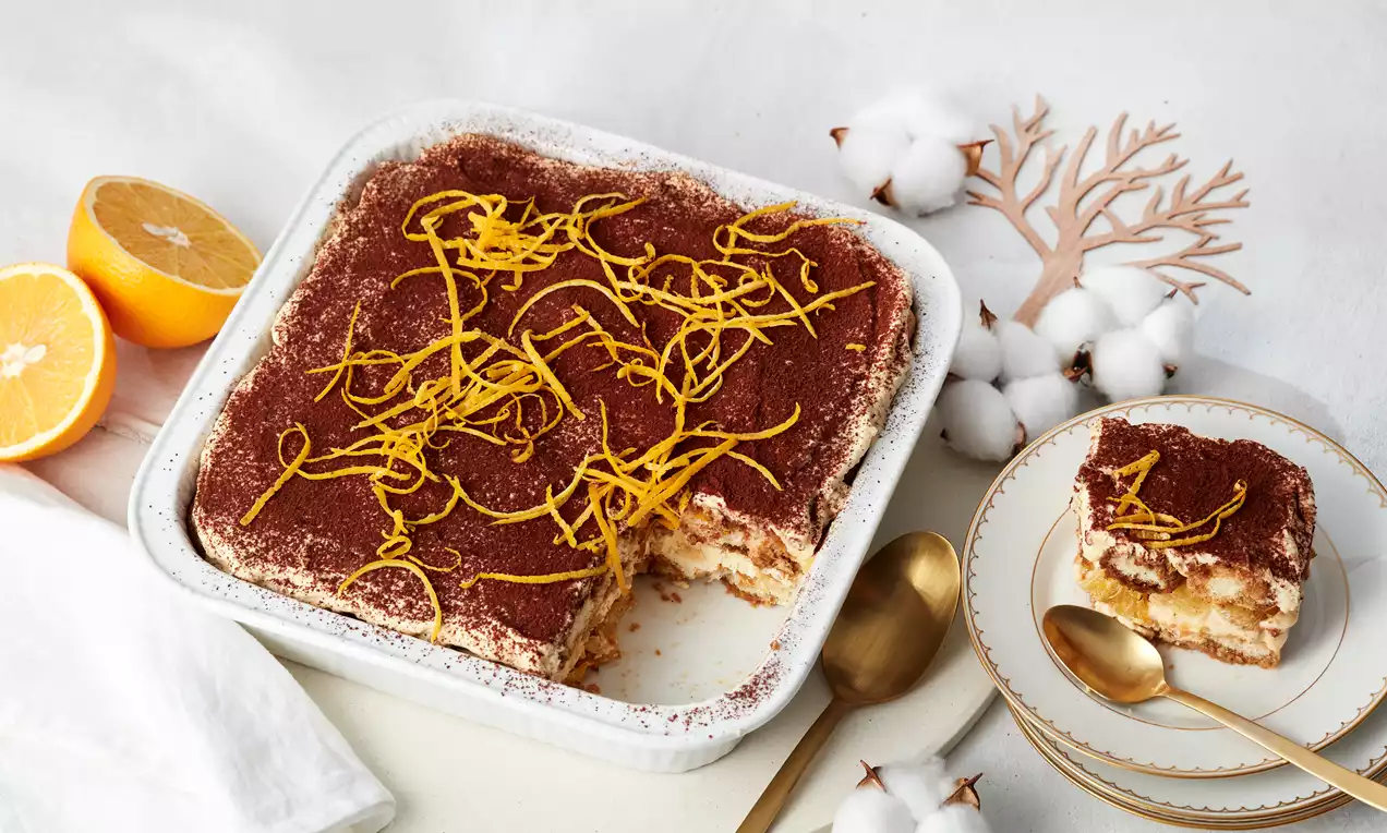 Tiramisu à l'orange présenté dans un bol blanc sur une table recouverte d'une nappe blanche. À côté, une part est déjà disposée sur une assiette.