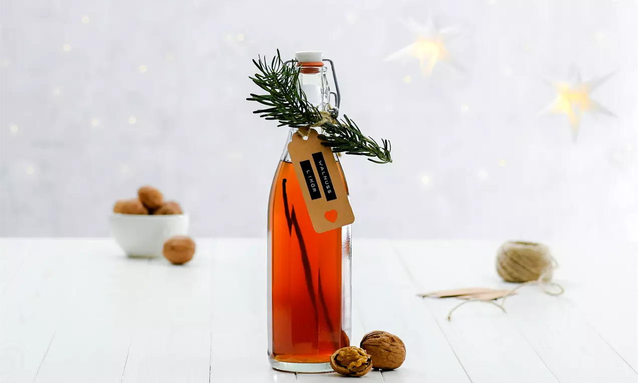 Schlanke Glasflasche mit bernsteinfarbenem Walnusslikör, verziert mit Rosmarinzweig und Etikett, auf hellem Tisch mit Walnüssen und Deko