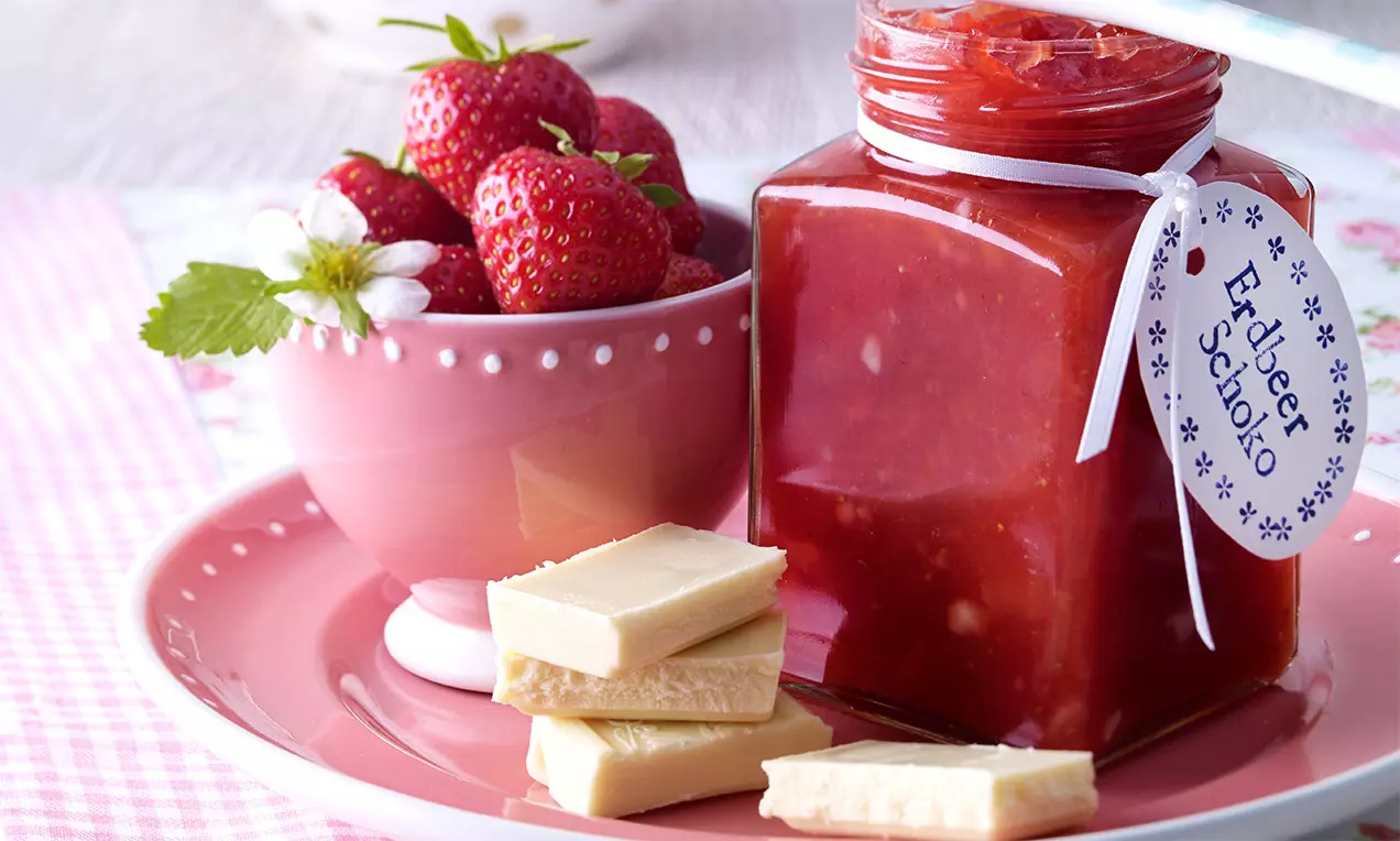 Confiture rouge vif de fraise dans un pot carré avec ruban blanc, accompagnée de fraises fraîches et carrés de chocolat blanc sur une assiette rose