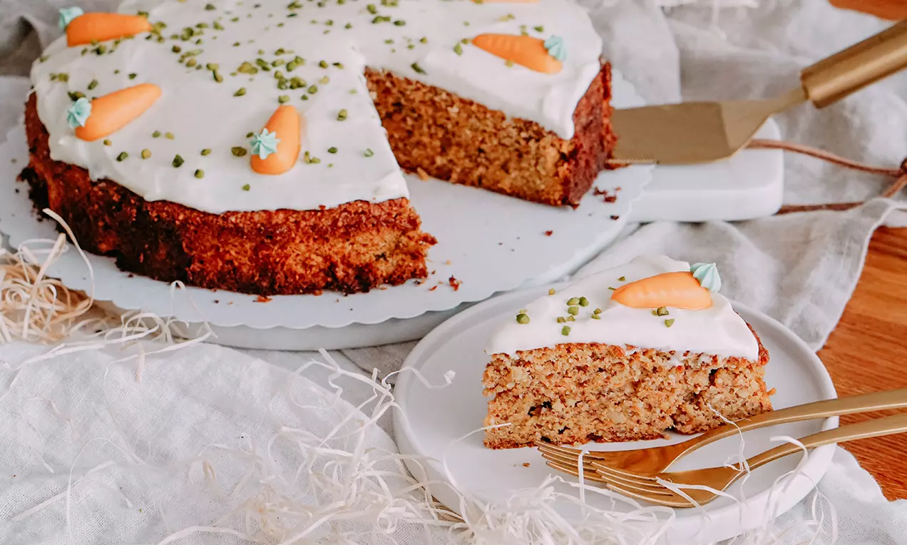 Glutenfreie Rüeblitorte mit saftigem, braunem Boden, weißer Glasur, Zuckermöhren und grünen Streuseln auf einem weißen Teller angerichtet