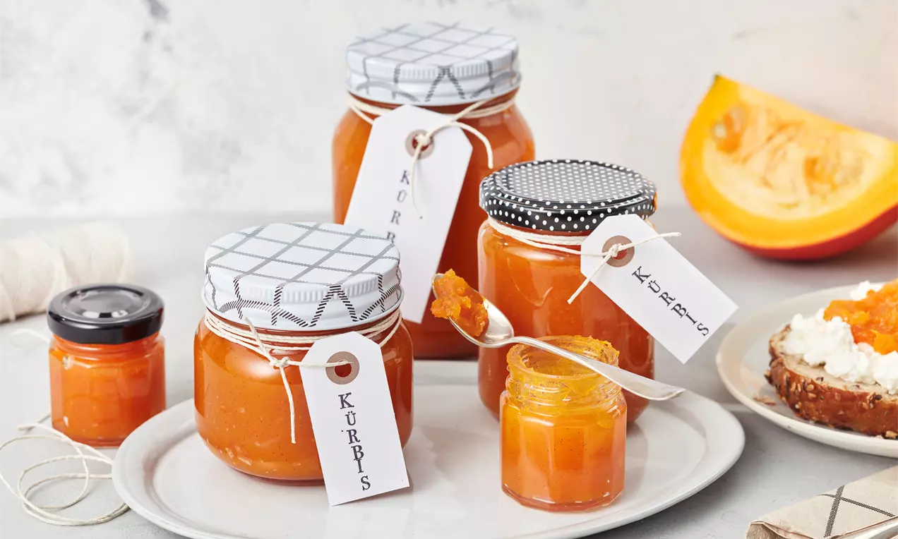 Glas mit leuchtend orangefarbener Kürbismarmelade, serviert mit einem Löffel auf einem weißen Teller neben Brot und Kürbisstücken