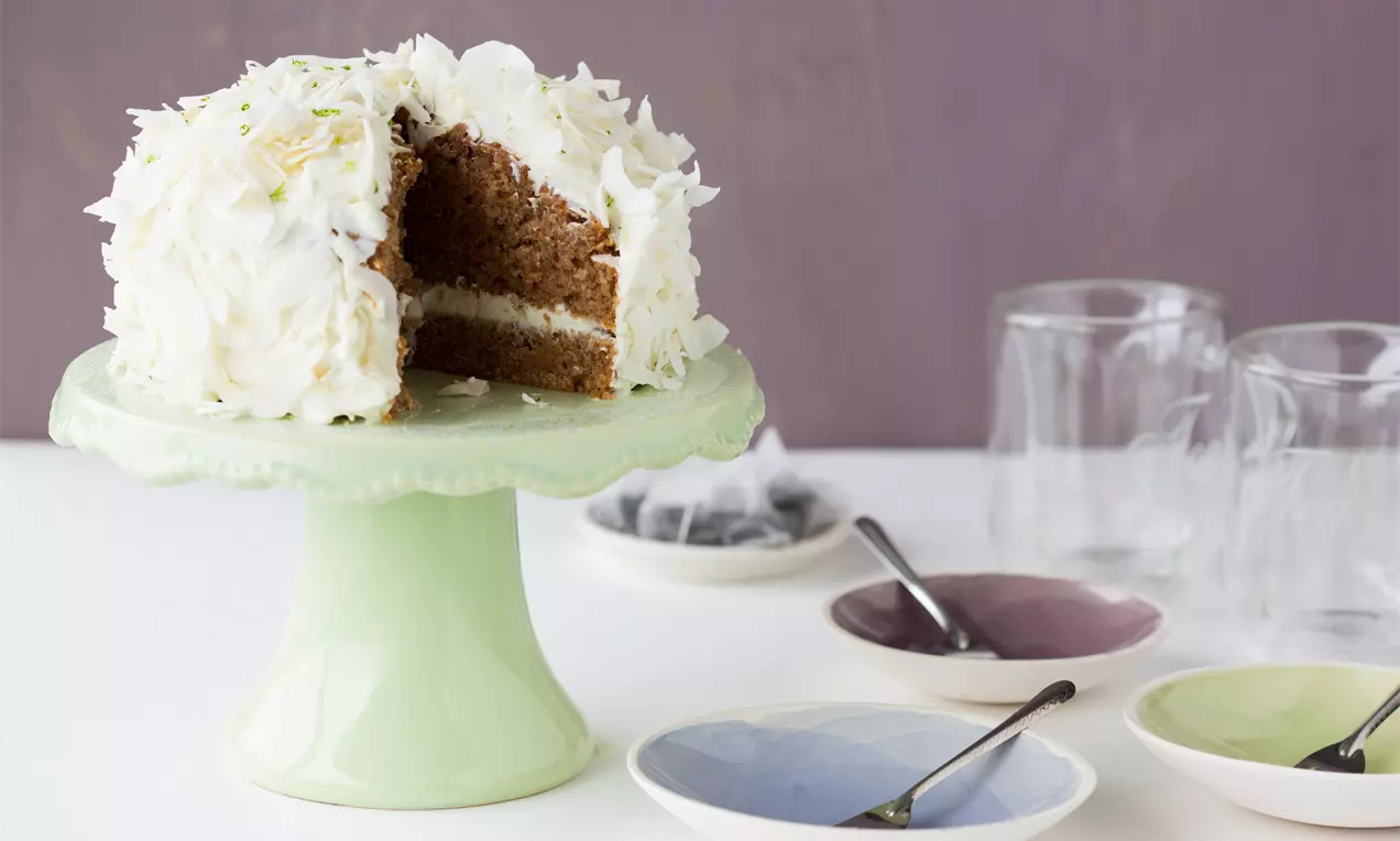 Rüebli-Limetten-Torte mit weißer Creme, dekoriert mit Kokosflocken und Limettenschale auf grünem Tortenständer, angeschnittene Stücke sichtbar