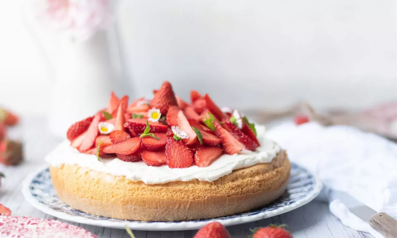 Locker gebackene Torte mit goldbraunem Boden, weißer Creme und frischen Erdbeerscheiben, dekoriert mit Blüten auf einem gemusterten Teller
