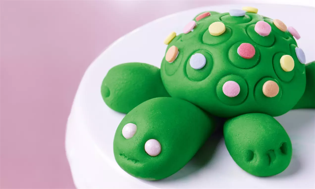 Grüne Marzipanschildkröte mit bunten Zuckerperlen auf dem gepunkteten Panzer, modelliert auf einer weißen Oberfläche mit rosa Hintergrund