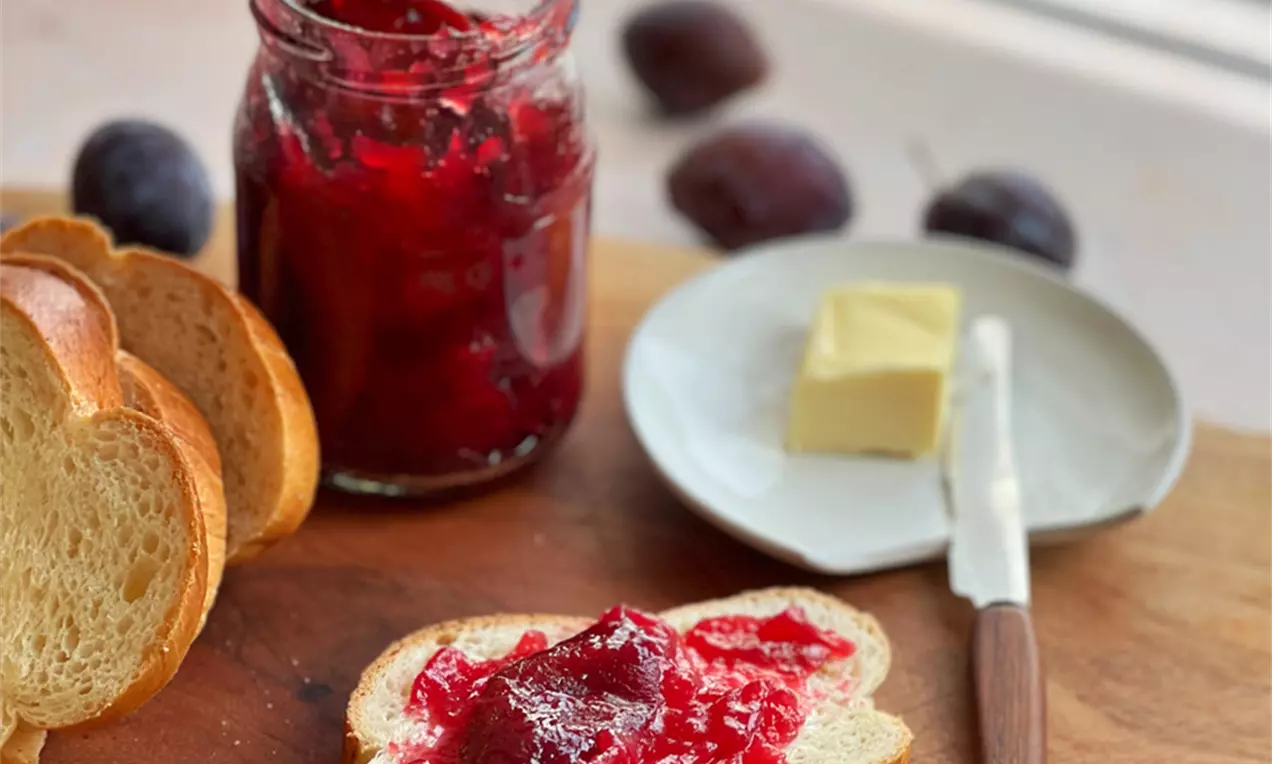 Ein Glas mit tiefroter Zwetschgenkonfitüre neben Butter und Brot auf einem Holzbrett, im Hintergrund frische Zwetschgen und sanftes Licht