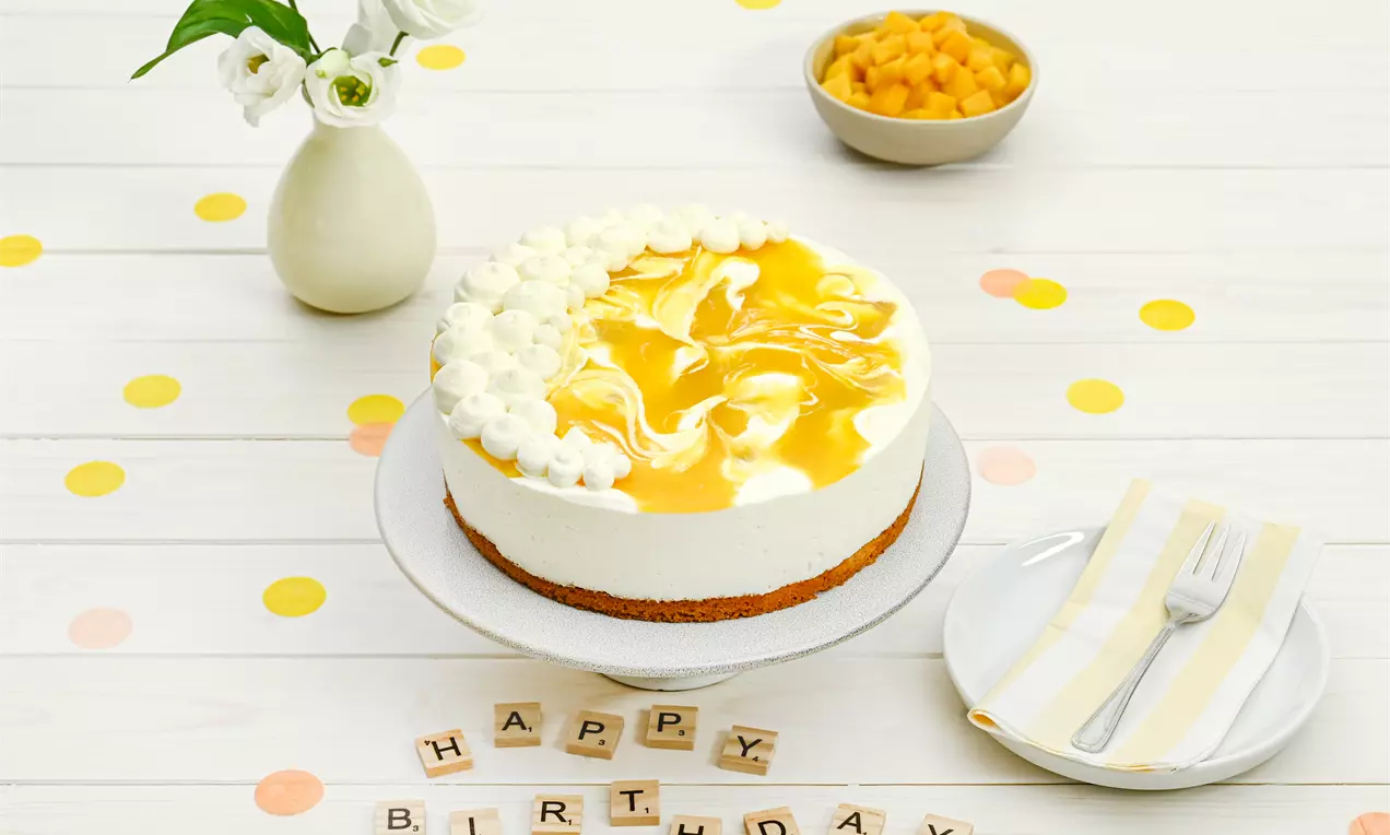 Käsesahne-Mango-Torte mit luftig weißer Creme, gelbem Mangospiegel und Sahnetupfen auf einer weißen Platte zwischen Geburtstagsdekoration
