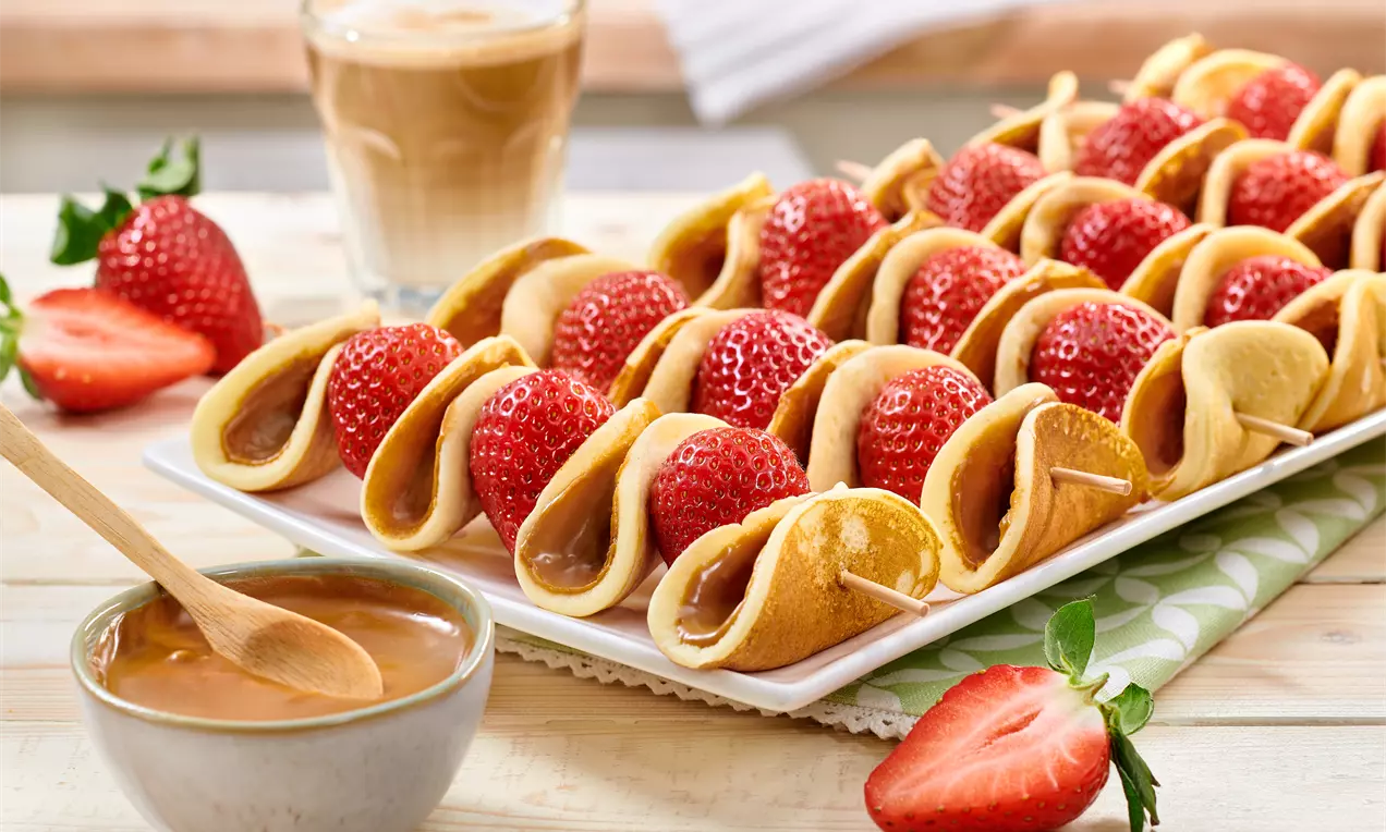 Spiedini con soffici pancake dorati piegati su fragole lucide rosso vivo, disposti ordinatamente su un vassoio bianco accanto a crema e caffè