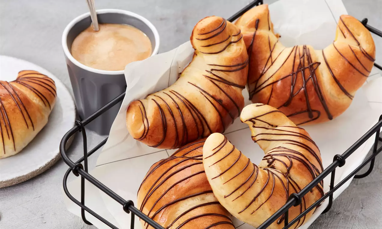 Goldbraune Schoko-Hörnchen mit geschwungener Form und dünnen Schokoladenstreifen auf weißem Backpapier in einem schwarzen Korb