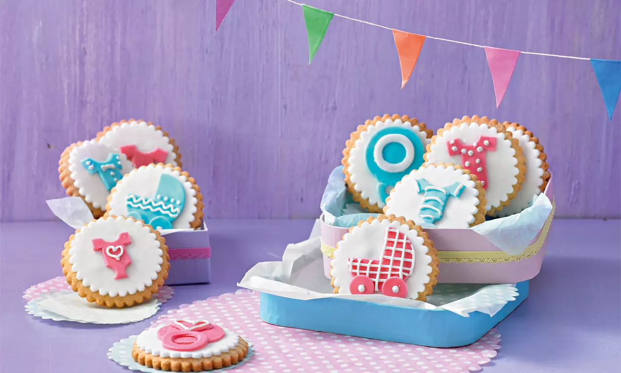 Runde Babykekse mit gezacktem Rand und bunter Zuckerdekoration auf weißem Fondant, angerichtet in einer pastellfarbenen Schachtel mit Papier ausgelegt