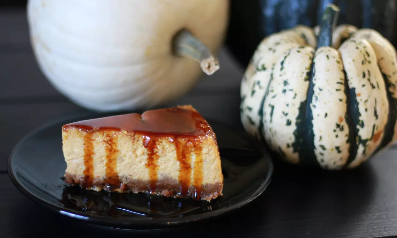 Part de cheesecake orange pâle sur une base foncée, nappée de caramel lisse, posée sur une assiette noire près de courges décoratives