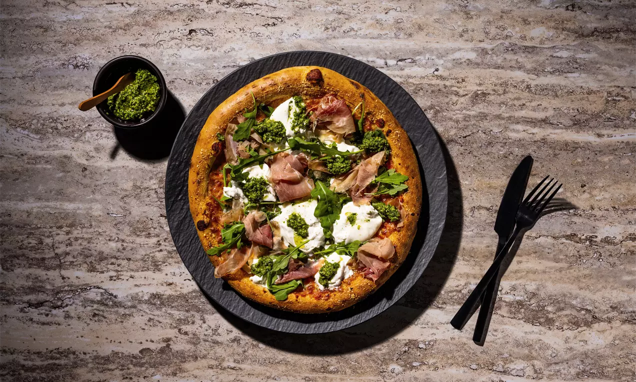 Pizza mit Parmaschinken und Burrata auf einer Suprema Margherita auf einem schwarzen Teller und marmoriertem Untergrund steht.