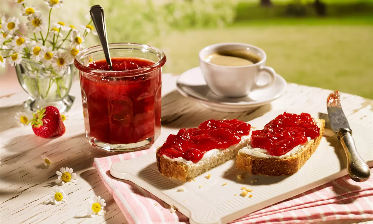 Erdbeerrote Konfitüre in Glas mit silbernem Löffel neben belegtem Brot auf weißer Platte, umgeben von Kamillenblüten und Espresso-Tasse