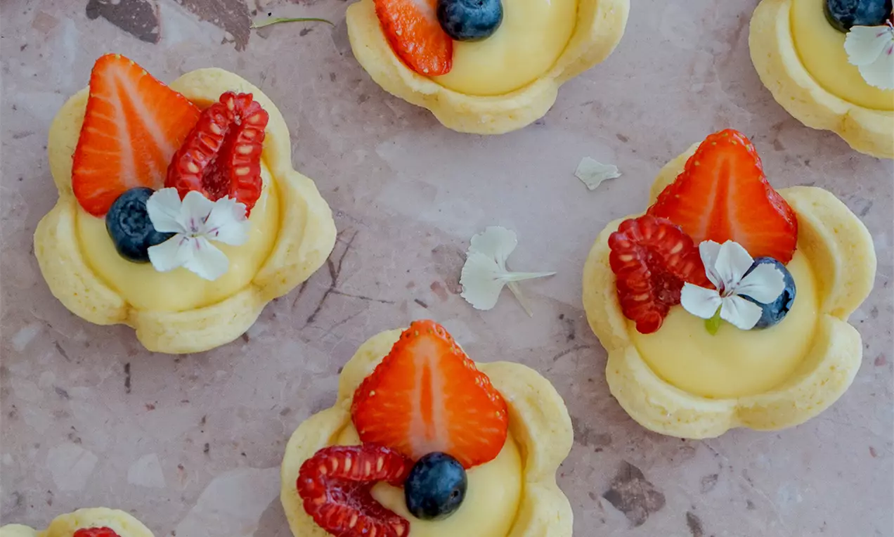 Butterzarte, blütenförmige Tartelettes mit gelber Zitronencreme, garniert mit Erdbeeren, Himbeeren, Blaubeeren und kleinen weißen Blüten