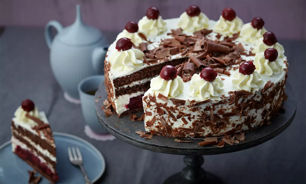 Schwarzwälder Kirschtorte mit dunklen Schokoraspeln, Sahnetupfen und Kirschen auf einer schwarzen Tortenplatte, daneben ein Stück Kuchen