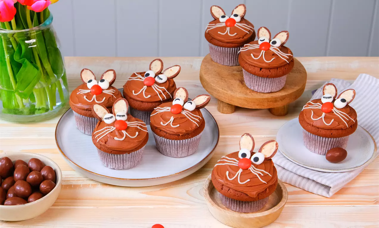 Muffins au chocolat brun surmontés de motifs de lapin avec oreilles en amandes, nez rouge et moustaches blanches sur assiettes et support bois