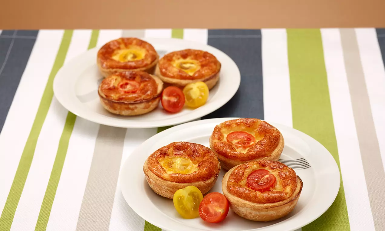 Piccole tortine salate dorate con guscio croccante e ripieno cremoso, guarnite con fette di pomodoro rosso e giallo su piatti bianchi