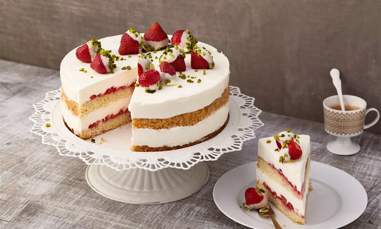 Torta con strati alternati di crema bianca e pan di Spagna dorato, decorata con fragole fresche e granella di pistacchio su un’alzata elegante