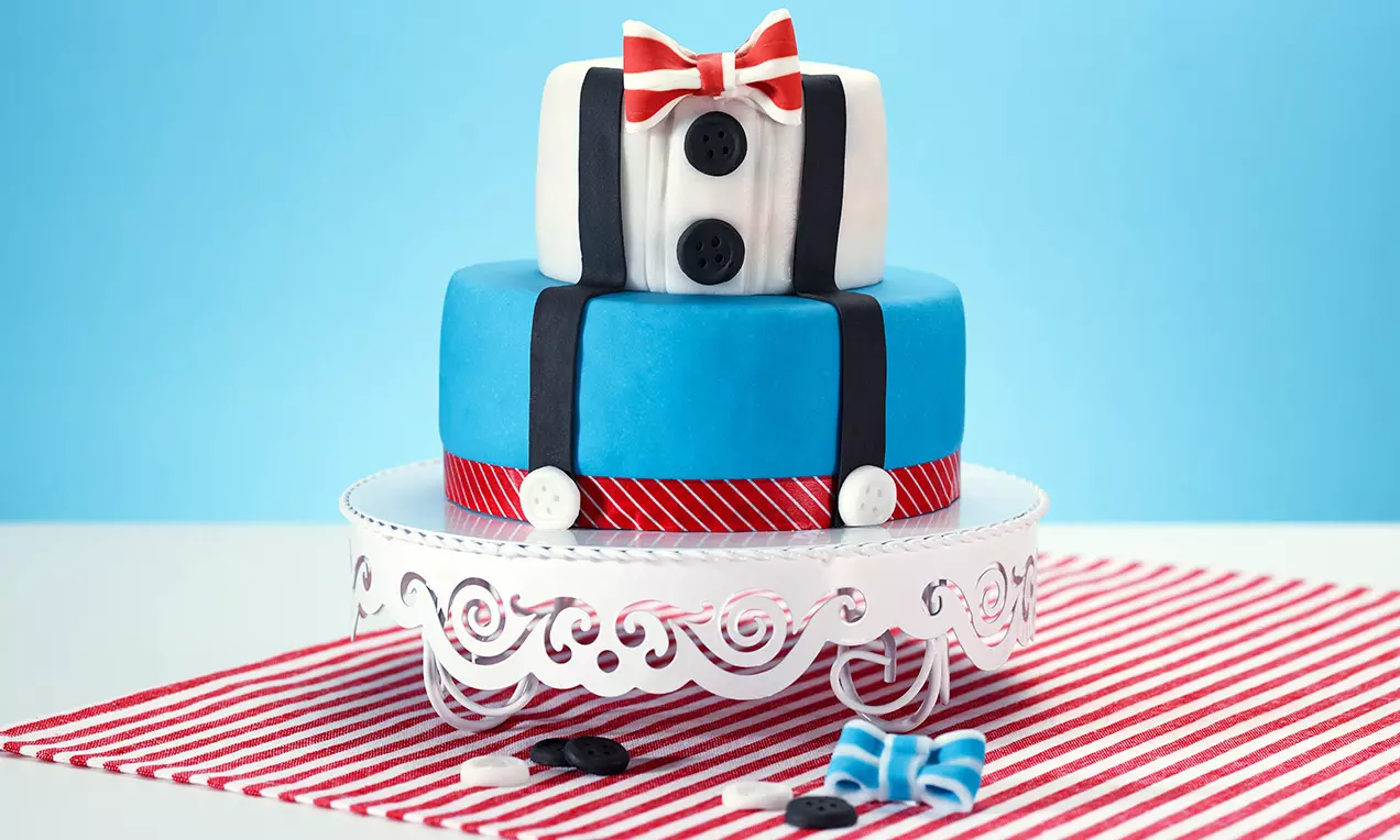 Gâteau en deux étages bleu et blanc décoré d'un nœud rouge, bretelles noires, boutons noirs et ruban rayé sur un support blanc ajouré