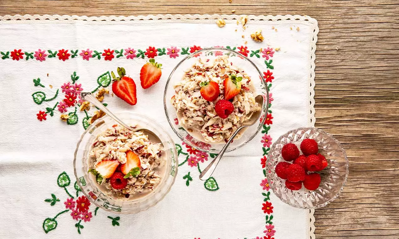 Fein geraffeltes Birchermüesli mit zarten Farben und fruchtigen roten Erdbeeren und Himbeeren in Glasdessertschalen auf floral bestickter Tischdecke