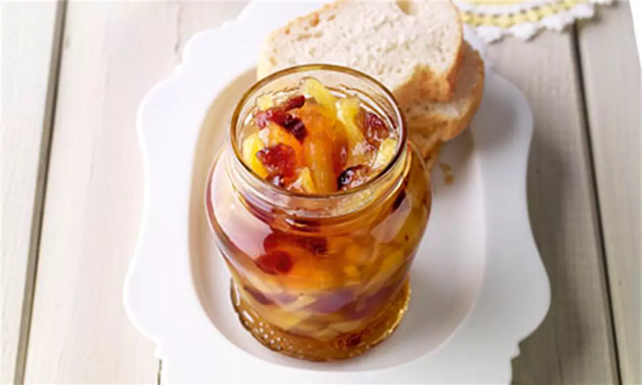 Goldgelbe Ananas-Cranberry-Konfitüre mit roten Fruchtstücken in einem Glas auf weißem Teller, dahinter Brotscheiben mit hellem Rand