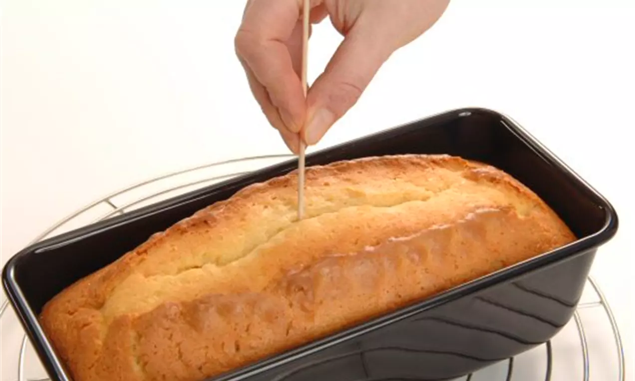 Hand hält ein Holzstäbchen, das in einen goldbraun gebackenen Kastenkuchen in schwarzer Backform auf einem silbernen Kuchengitter gesteckt wird