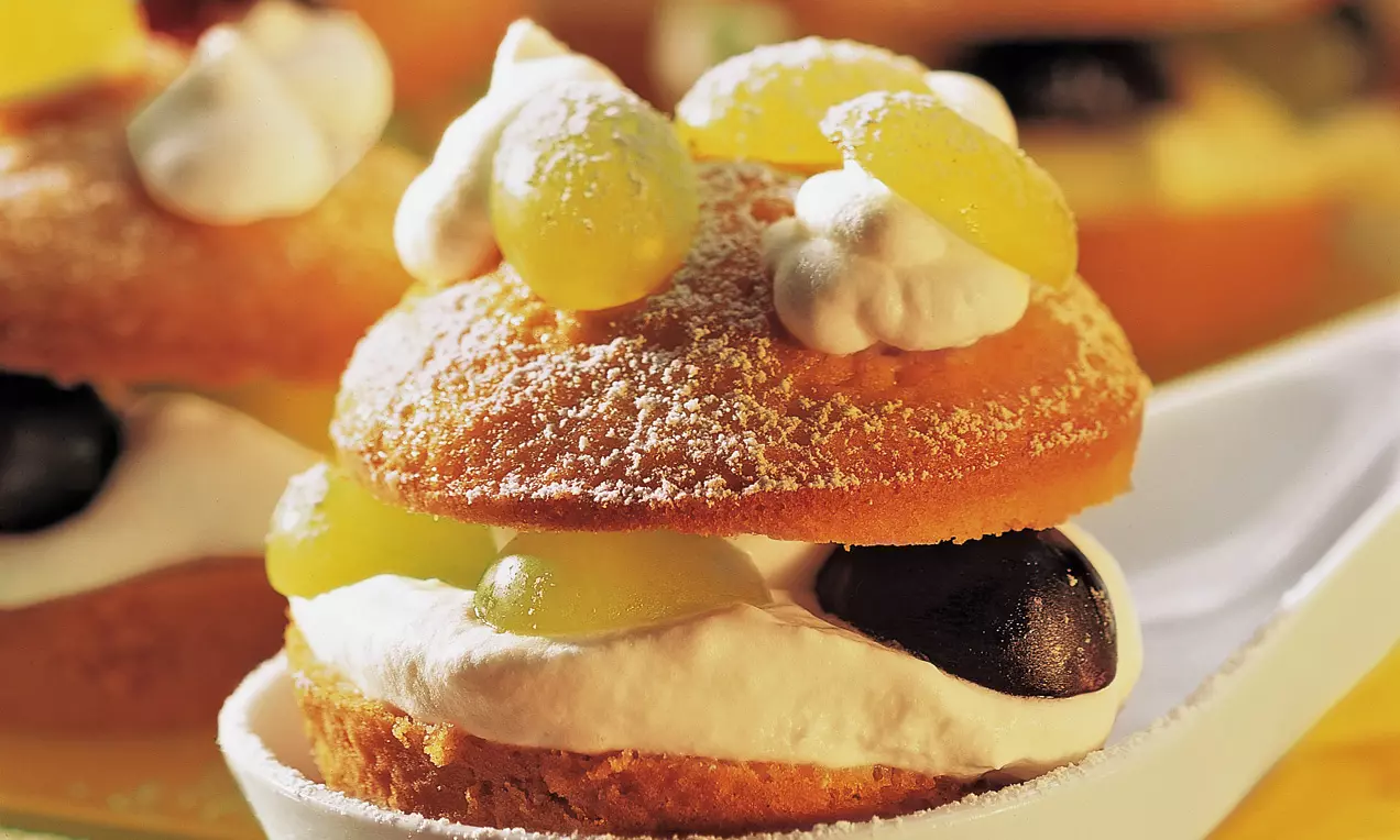 Muffin dorati ripieni di crema bianca soffice, decorati con uva gialla e nera, spolverati di zucchero a velo su un piatto chiaro