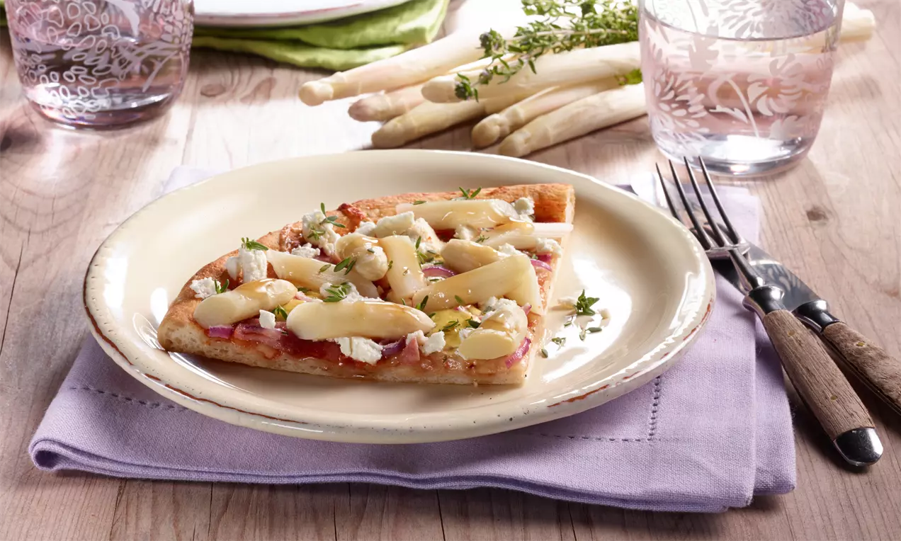 Ein Stück goldbraune Pizza Hawaii mit Spargel, cremigem Ziegen-Frischkäse und frischen Kräutern auf einem beigen Teller, dekoriert mit Flieder-Serviette