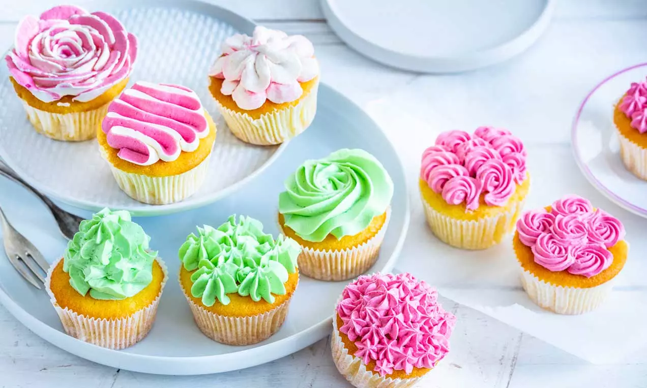 Cupcake dorati con morbida glassa colorata verde, rosa e bianca, decorati a rose e swirl, disposti su piatti chiari su sfondo bianco