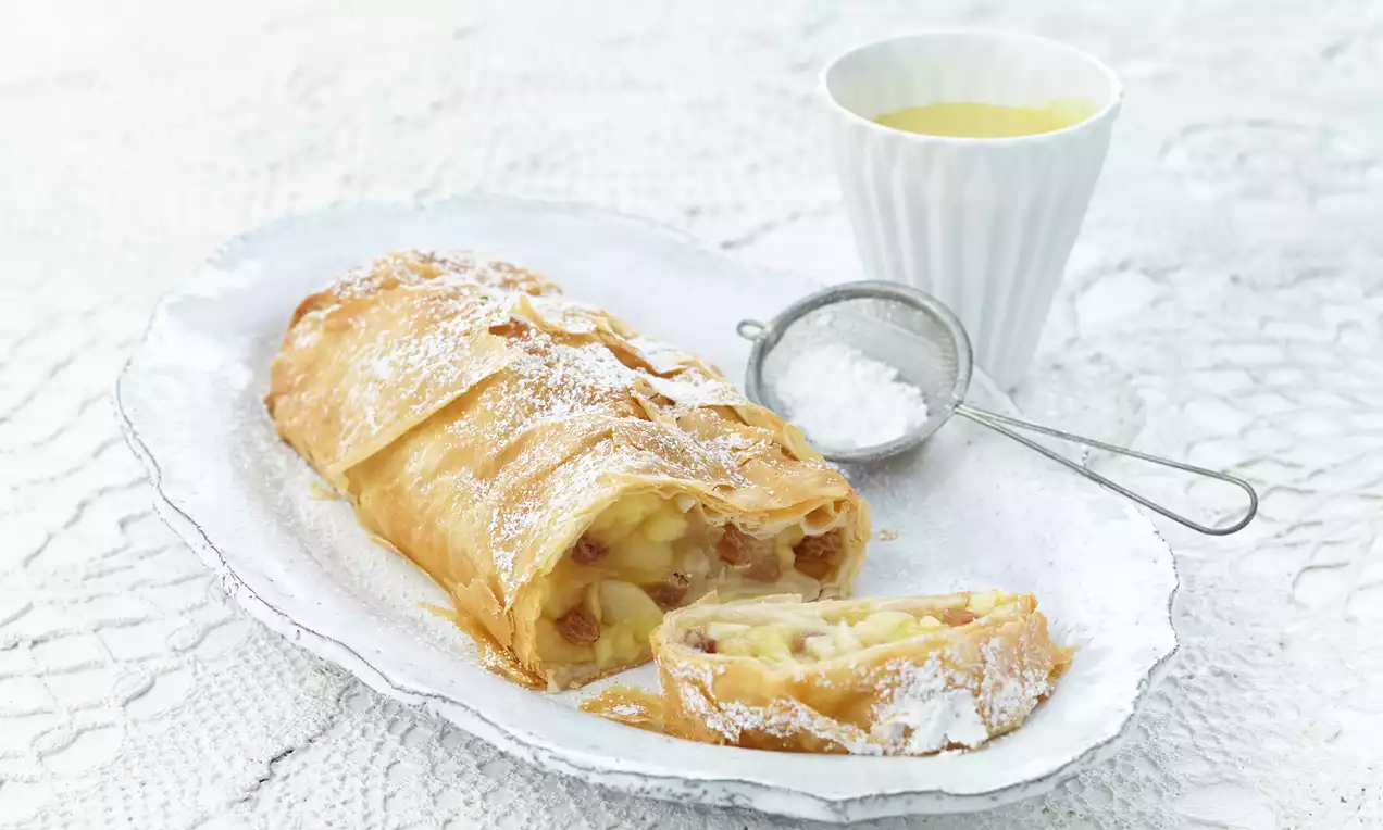 Goldbrauner Wiener Apfelstrudel mit zartem Blätterteig und saftiger Füllung aus Äpfeln, Rosinen und Zimt auf einem weißen Teller mit Puderzucker-Sieb