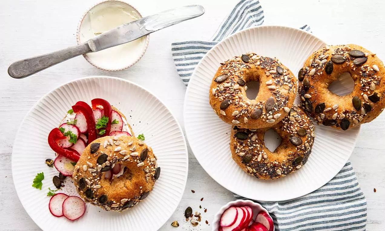Vollkorn-Bagels mit dunkler Kruste und vielen Körnern auf weißen Tellern, dazu Frischkäse, Radieschenscheiben und gestreiftes Tuch drapiert