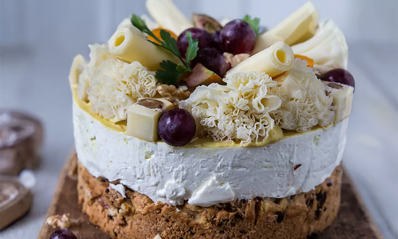 Salziger Cheesecake mit weißer Frischkäsefüllung auf goldbraunem Nussboden, dekoriert mit Trauben, Käserollen, Nüssen und Kräutern