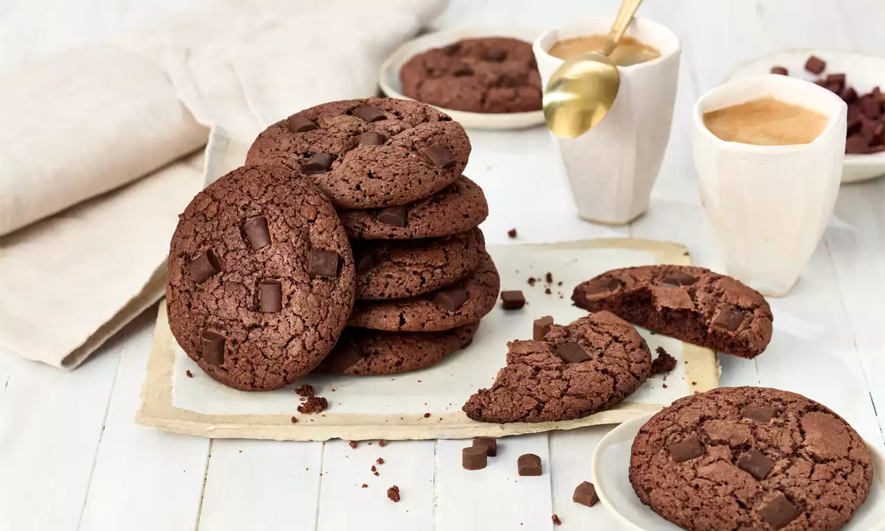 Schokoladenbraune, weiche Brownie-Cookies mit dunklen Schokostückchen, teils zerbrochen, auf einem rustikalen Teller neben Kaffee-Tassen arrangiert