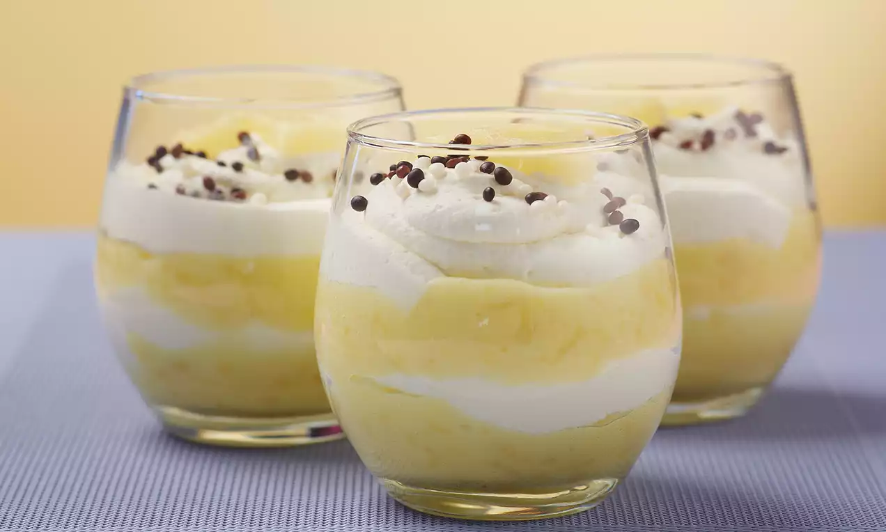 Schichtdessert aus gelber Ananascreme und weißer Sahne im bauchigen Glas, oben mit dunklen Schokostückchen bestreut auf lilafarbenem Untergrund