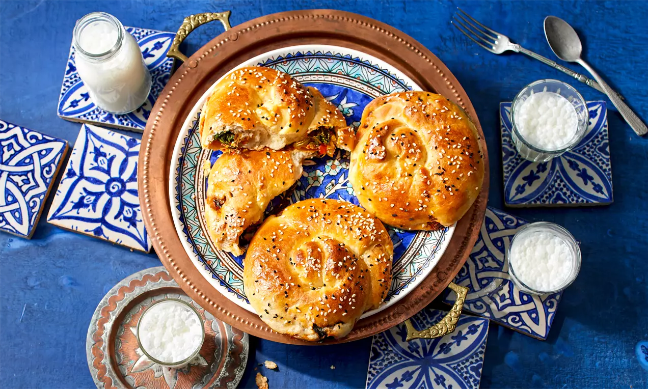 Goldbraun gebackener Börek mit Spinatfüllung, spiralförmig mit Sesam bestreut auf verziertem Keramikteller, umgeben von traditionellen Mustern
