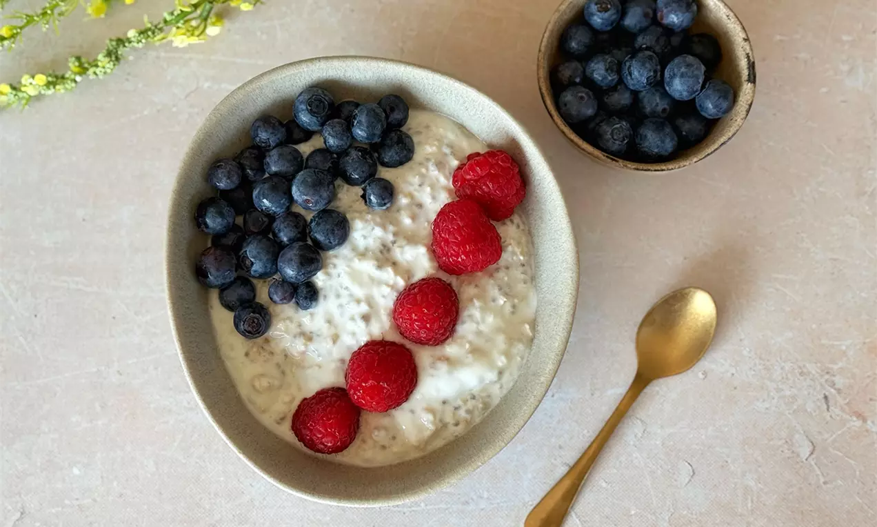 Cremiges Birchermüesli in beigem Teller mit frischen Blaubeeren und roten Himbeeren garniert, daneben goldener Löffel und Schale Blaubeeren