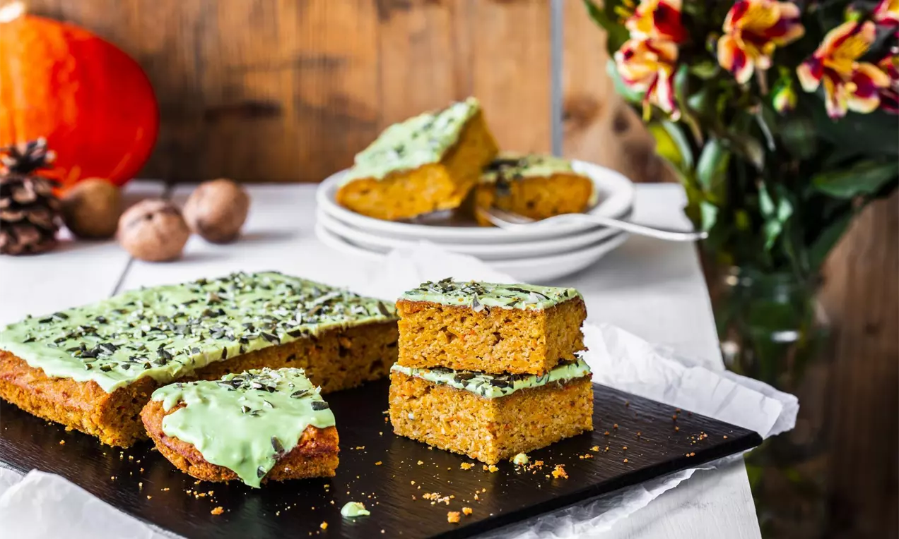 Torta rettangolare di zucca con glassa verde chiaro e granella di cioccolato scuro su un vassoio nero, accanto a piatti bianchi e decorazioni autunnali