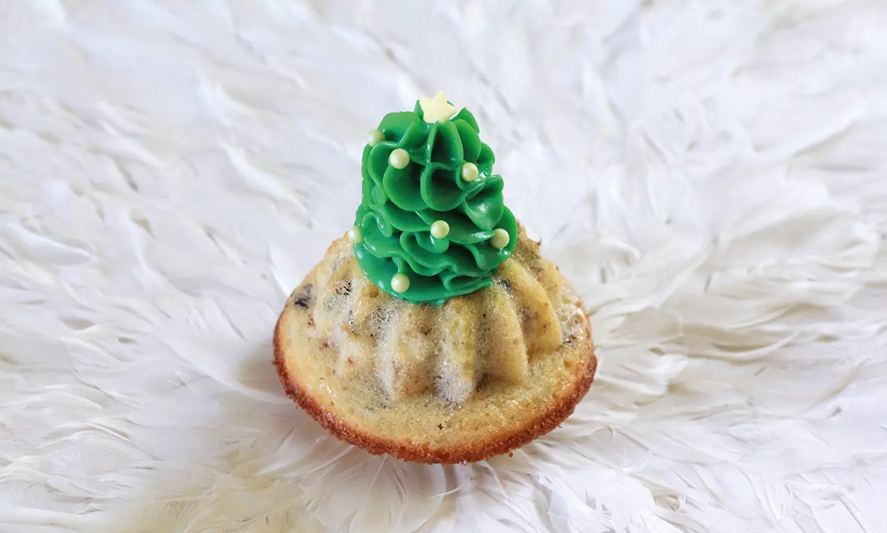 Ein goldbrauner Mini-Gugelhupf mit grüner Tannenbaumhaube aus sahniger Creme und weißen Perlen auf strukturiertem weißem Hintergrund
