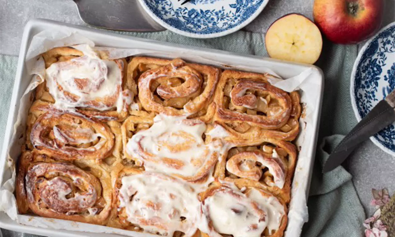 Goldbraune Cinnamon Rolls mit glänzender Zuckerglasur in weißer Backform, umgeben von frischen Äpfeln und blau gemusterten Tellern