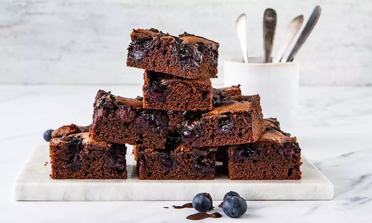Brownies moelleux couleur chocolat profond avec des myrtilles juteuses incrustées, disposés en pile sur une planche marbrée blanche