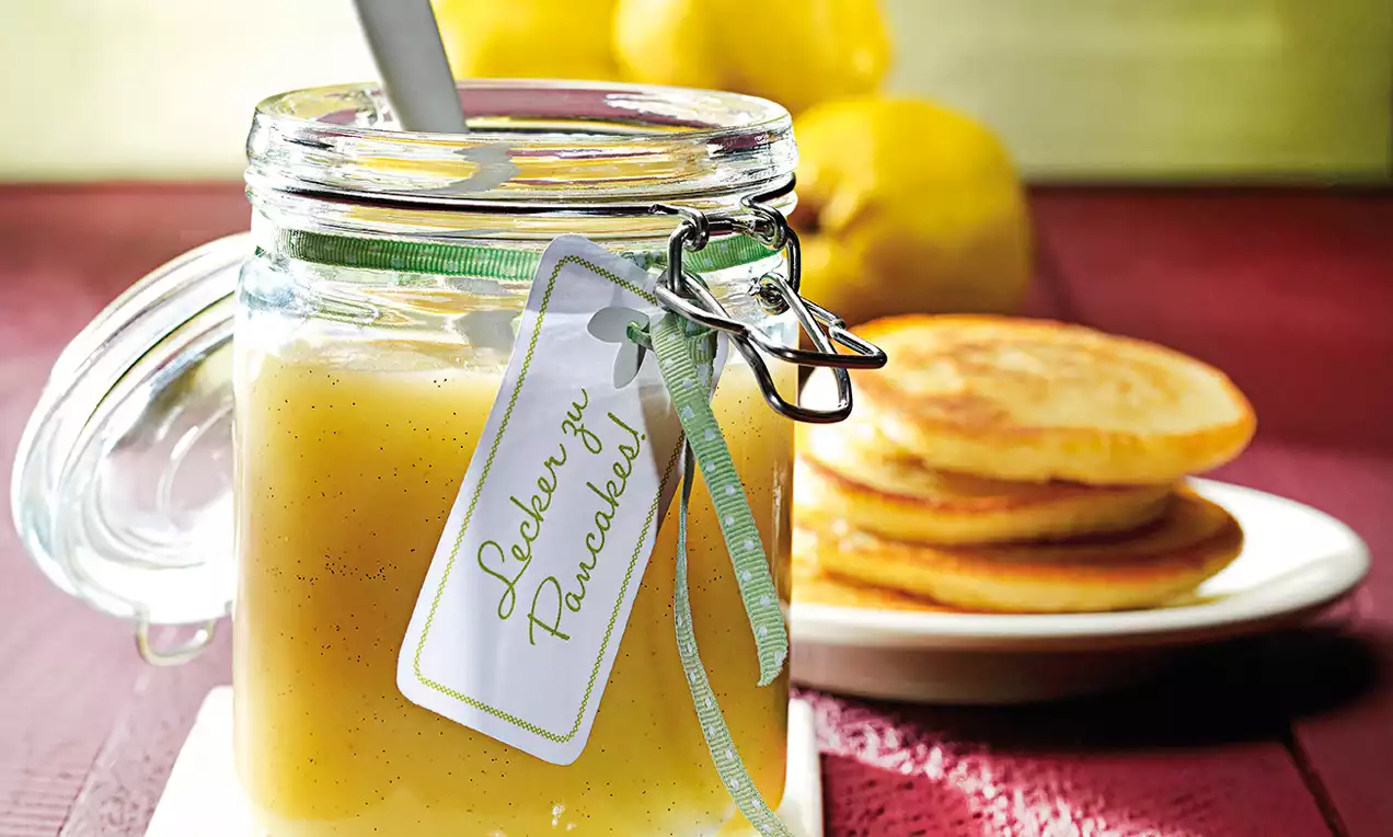 Im Glas mit Bügelverschluss Quitten-Apfel-Mus in zartem Gelb, cremig und glatt, daneben gestapelte goldbraune Pancakes auf weißem Teller