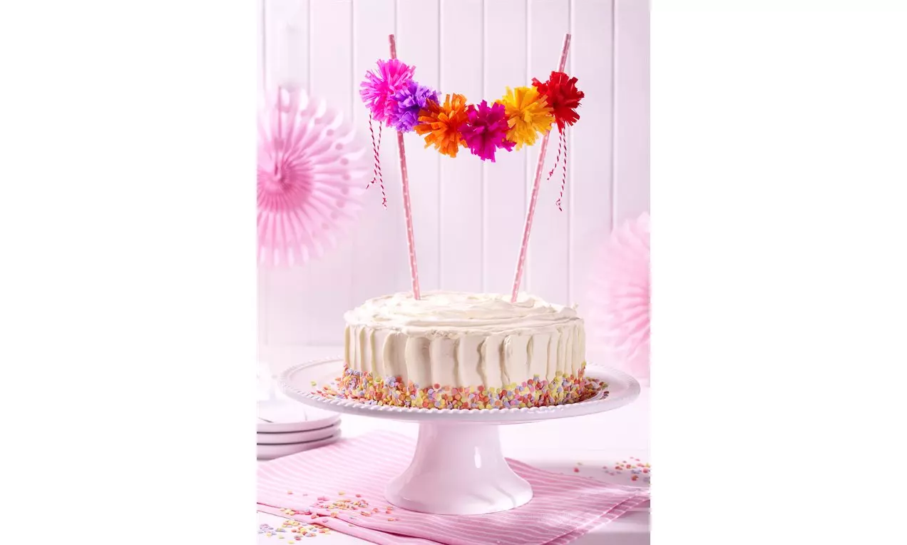 Weiße Torte mit glatter Creme, verziert mit bunten Zuckerstreuseln und mehrfarbigen Pompons an Stäben auf einer weißen Tortenplatte