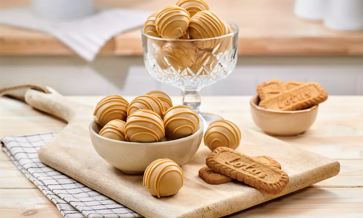 Tartufi dorati dal rivestimento lucido con linee di crema caramello, disposti in ciotole di ceramica e una coppa di vetro accanto a biscotti Lotus