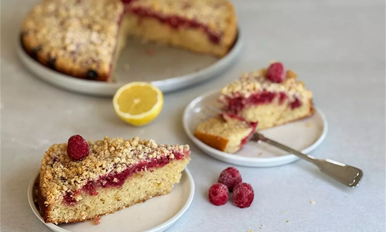Zitronen Himbeer Streuselkuchen mit goldgelbem Boden, roter Himbeerschicht, knusprigen Streuseln und frischen Himbeeren auf weißen Tellern