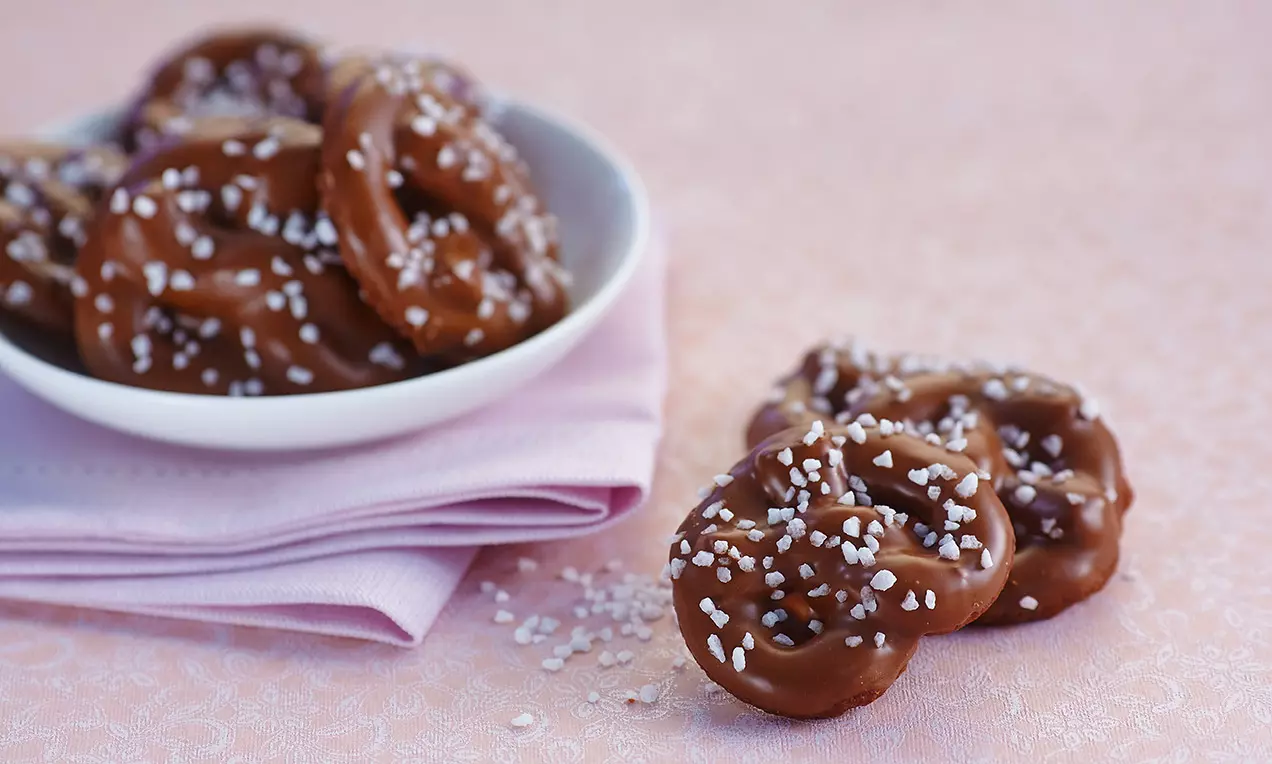 Bretzels enrobés de chocolat brillant garnis de grains de sucre blanc, disposés sur une serviette rose et un fond clair