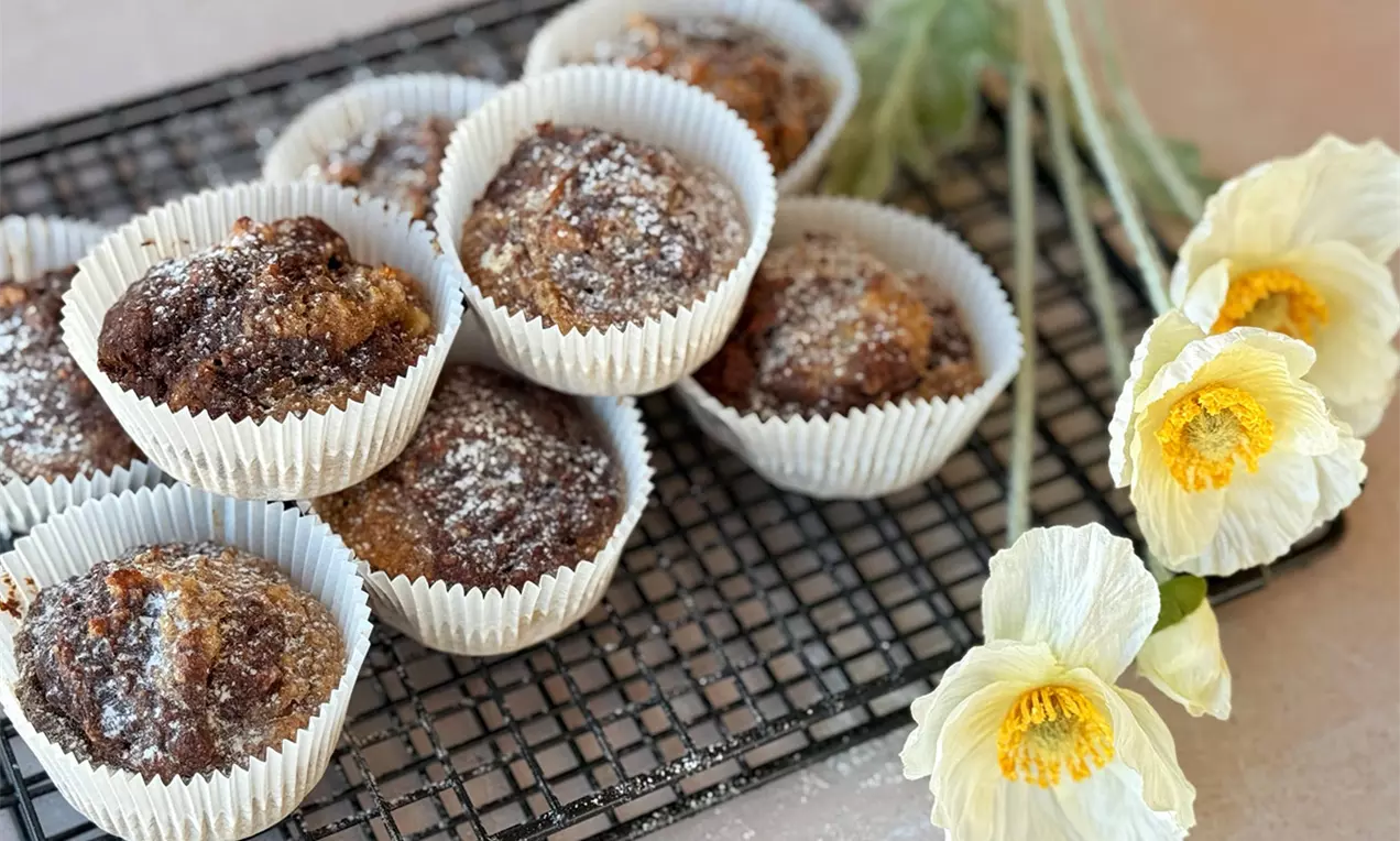 Zuckerfreie Marmor-Bananen-Muffins mit goldbrauner Oberfläche und leichter Puderzucker-Schicht in weißen Förmchen auf schwarzem Gitter neben gelben Blumen